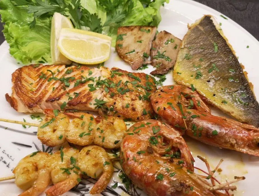 Piatto di frutti di mare grigliati con salmone, gamberi, pesce, spicchio di limone e verdure a foglia verde su un piatto bianco.