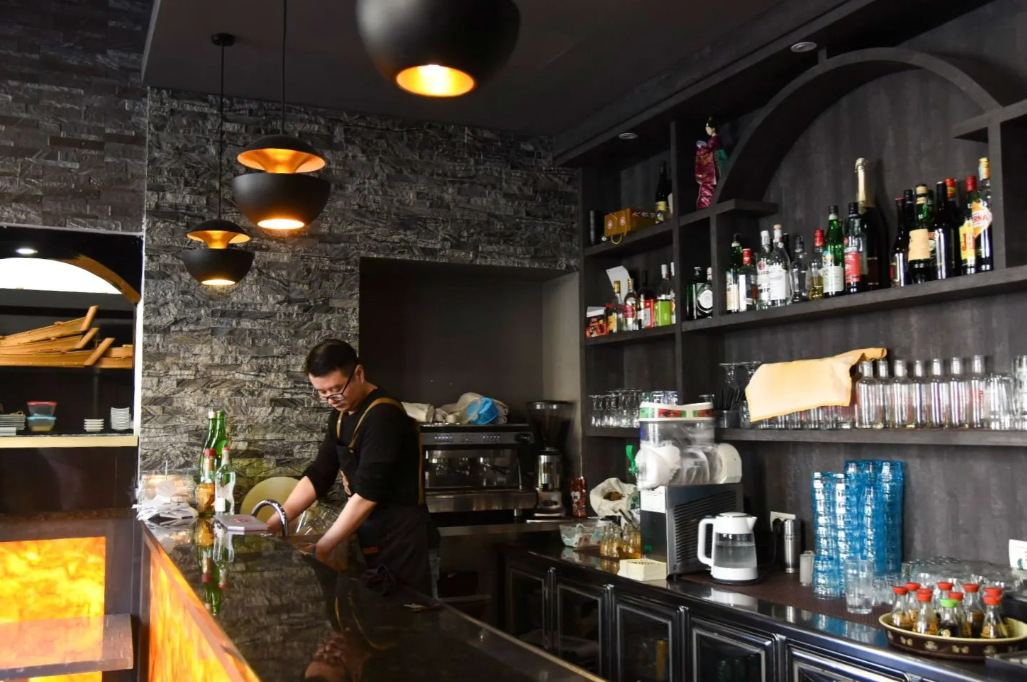Un barista lavora dietro un bancone scarsamente illuminato, con scaffali pieni di bottiglie e bicchieri, e una fiamma arancione che brilla sulla sinistra.