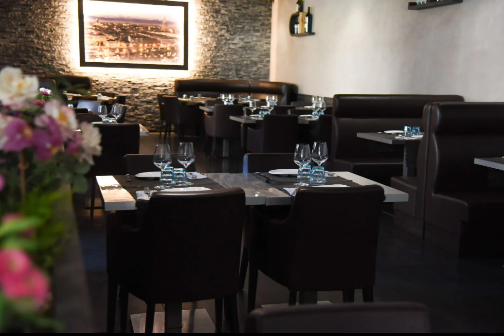 Sala da pranzo di un ristorante scarsamente illuminata con tavoli neri, bicchieri disposti con cura e una composizione floreale in primo piano.