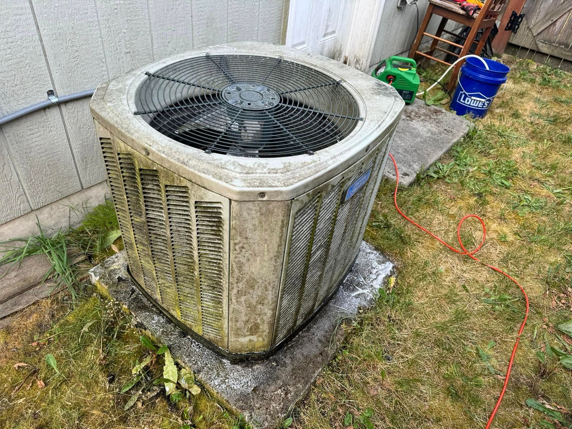 Outdoor air conditioner unit beside a wall, with a red extension cord on the grass.
