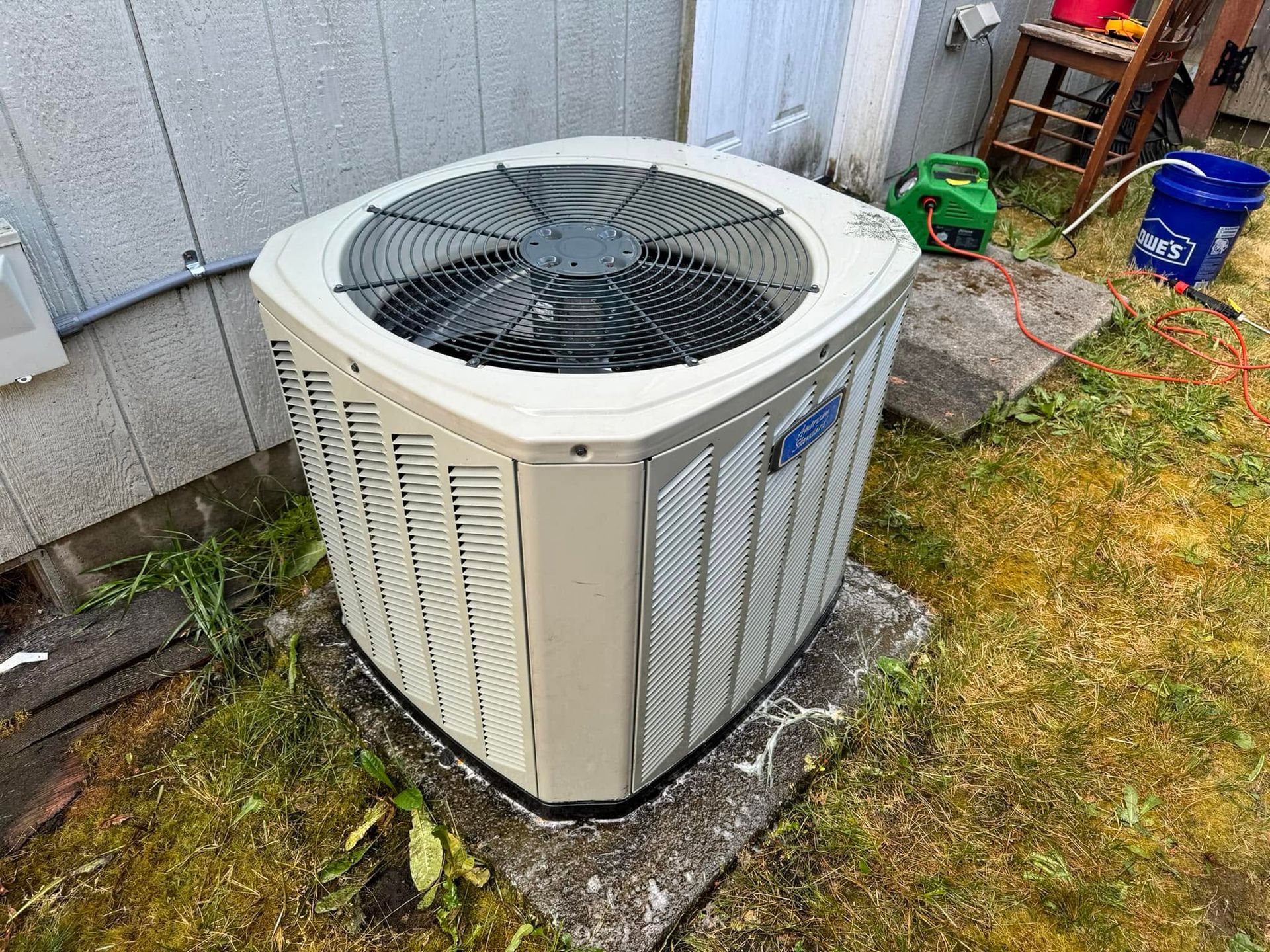 Outdoor air conditioner unit beside a house, on a concrete pad with grass and utility items nearby