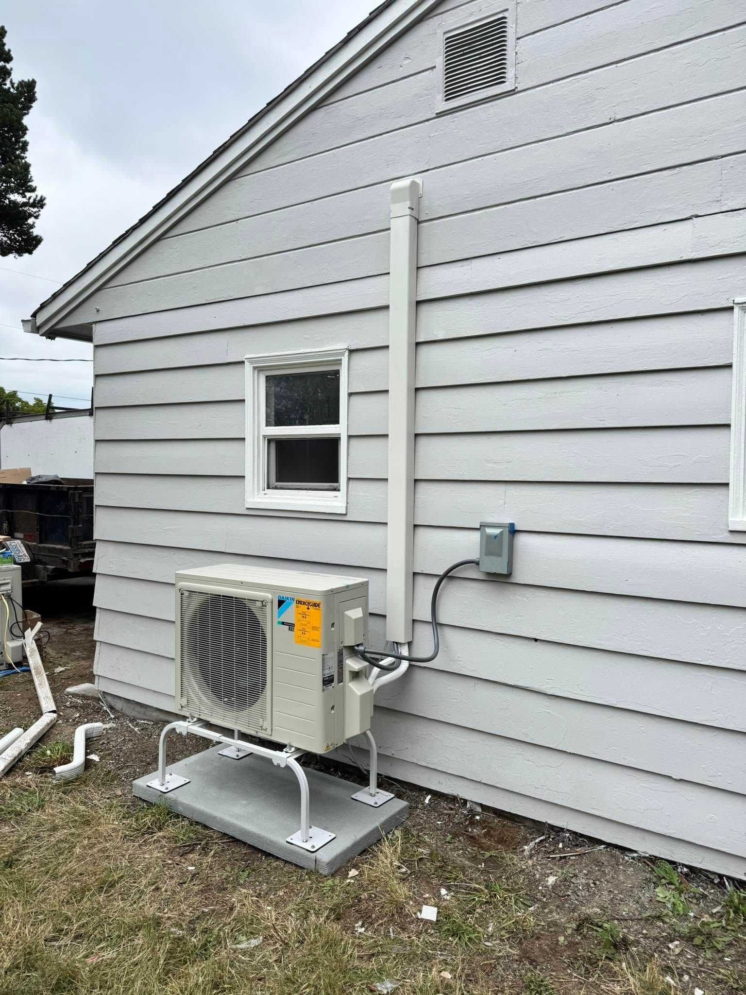 Side of a gray house with an outdoor HVAC unit and utility conduit beside a small window