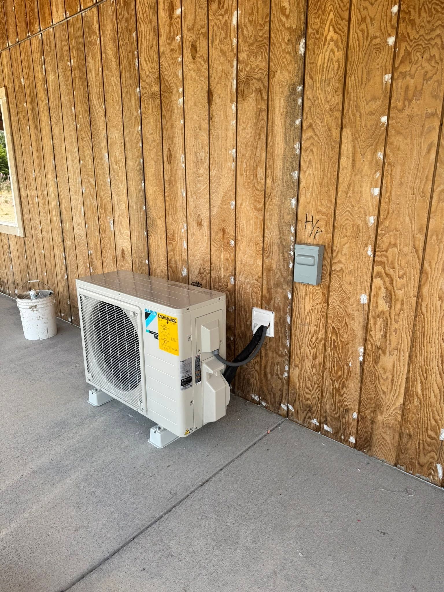 Outdoor AC unit beside a wooden wall, with a plugged-in power cord on a concrete floor.