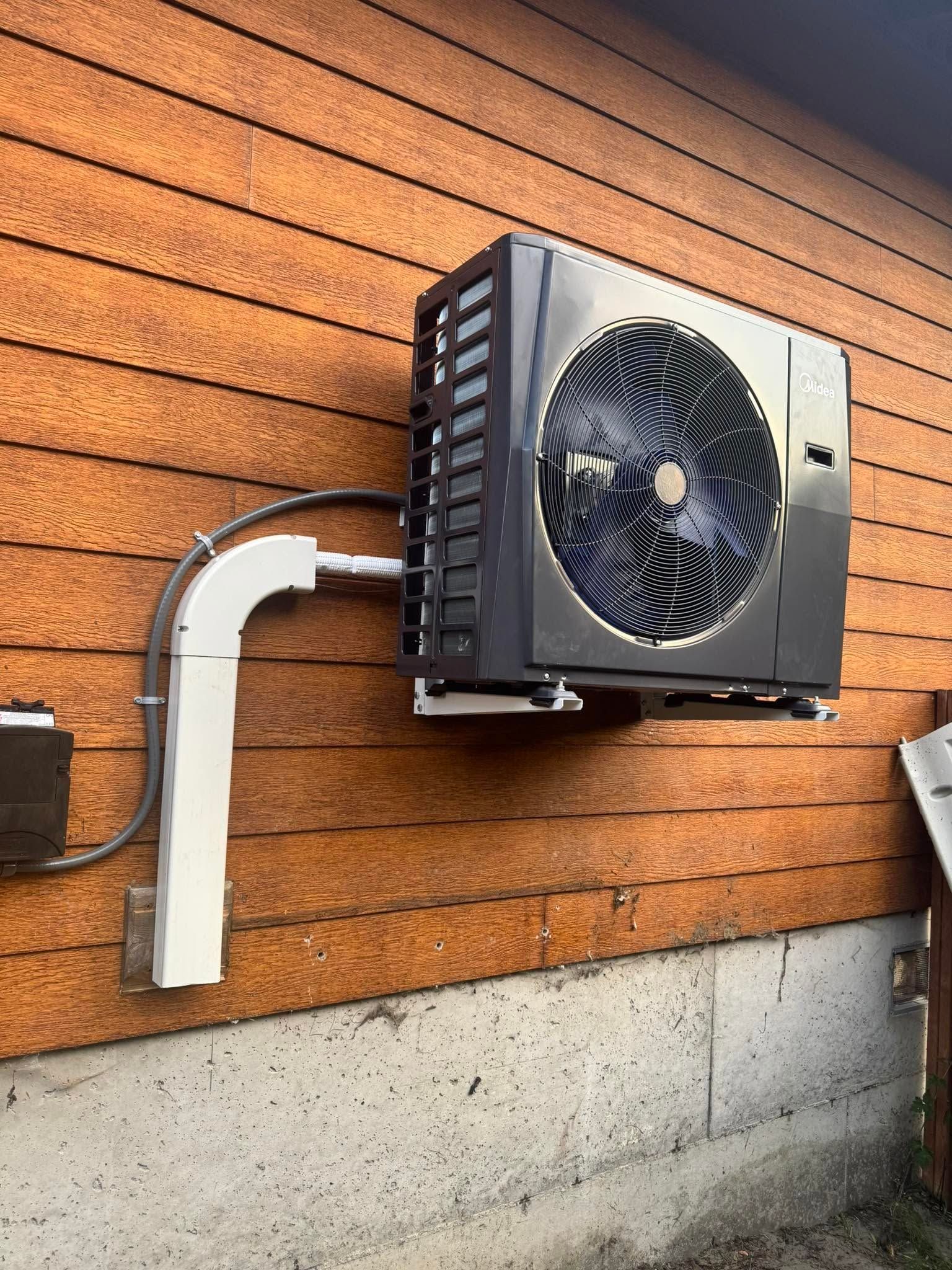 Outdoor wall-mounted air conditioner unit on a wooden-sided building with a white conduit and concrete base.