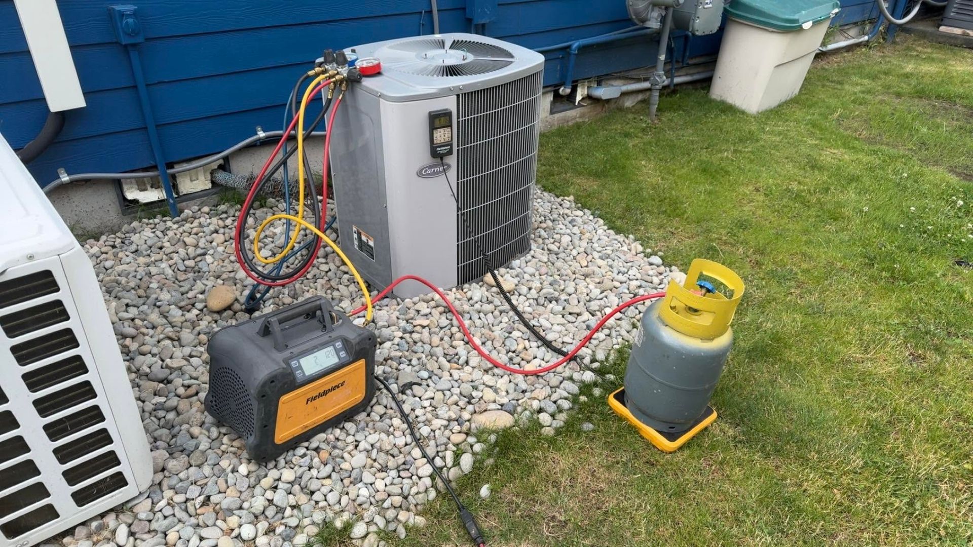 Outdoor HVAC unit with gauges and hoses connected to a small yellow tank on gravel beside a house