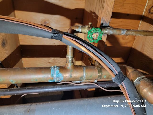 Copper pipes with green-handled valve, black insulation, and signs of corrosion in a wooden setting.
