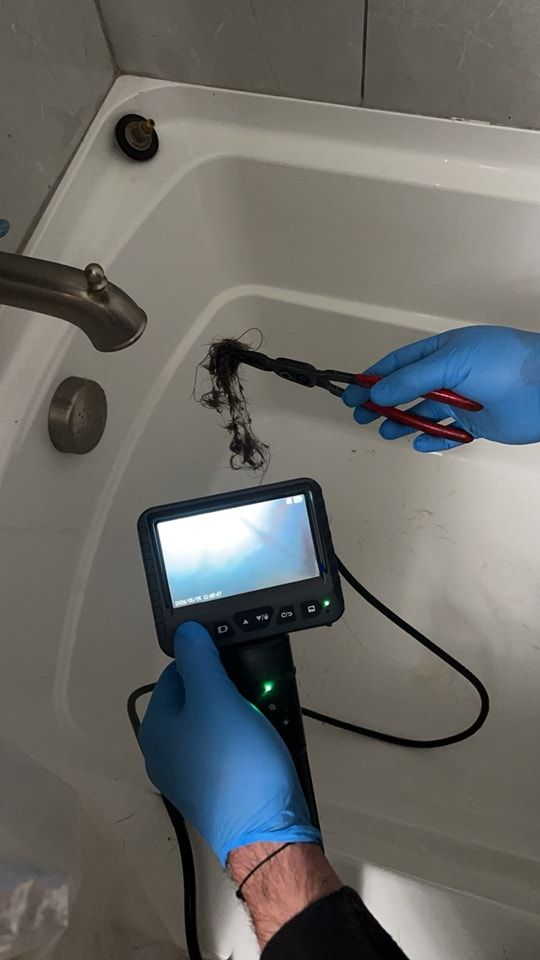 Person with gloved hands using pliers to remove hair from a bathtub drain, with a drain inspection camera.