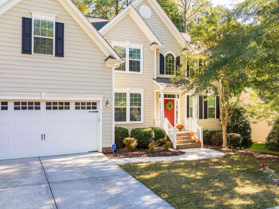 Two-story beige suburban house with white garage, red front door, and a sunlit driveway.