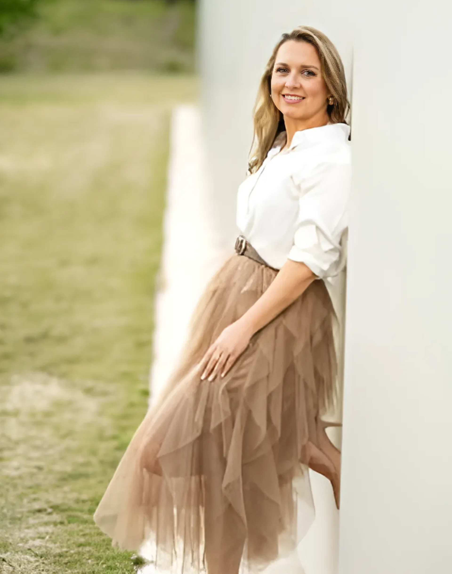 Woman leans against white wall, smiling. She wears a white shirt, brown tulle skirt, and belt; outdoor setting.