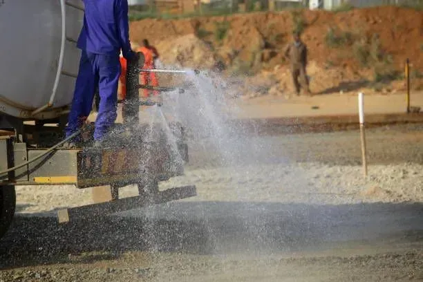 A person in blue work clothes sprays water from a construction tanker onto a dirt road at a job site.