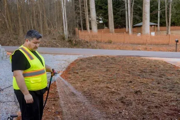 A person wearing a high-visibility vest uses a hose to spray water onto a patch of dirt next to a wooden fence.