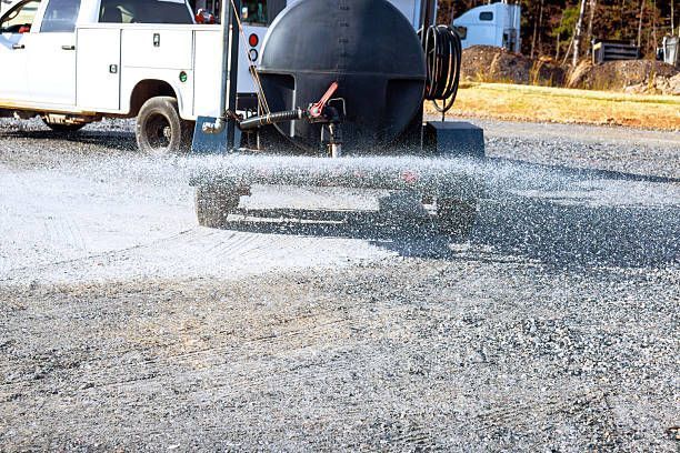 A white utility truck towing a tank trailer sprays a liquid onto a gravel surface.