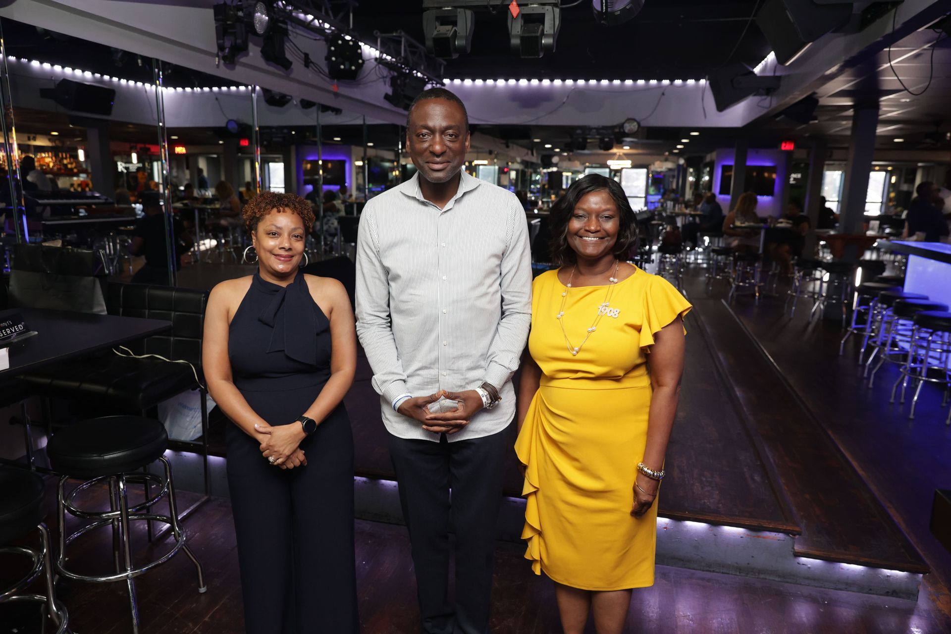 Three people stand inside a venue. Woman in yellow dress, man in button-down shirt, woman in jumpsuit pose.