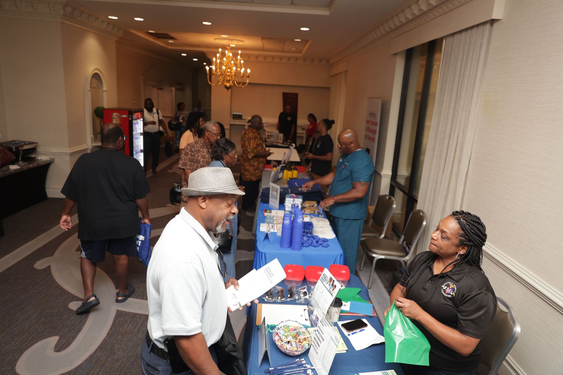 People at a health fair with tables, signage, and a chandelier in a well-lit room.