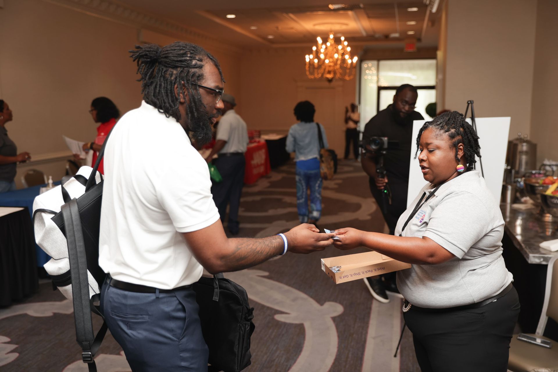 Man hands something to a woman at an event, with other people in the background.