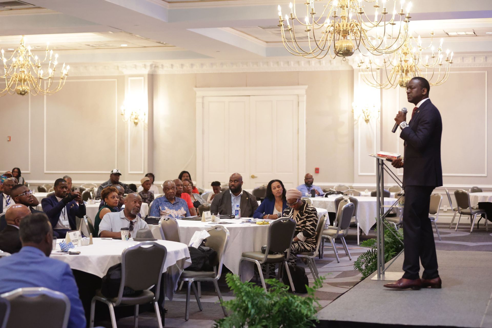 Man speaking at a conference. Attendees seated at round tables in a ballroom.