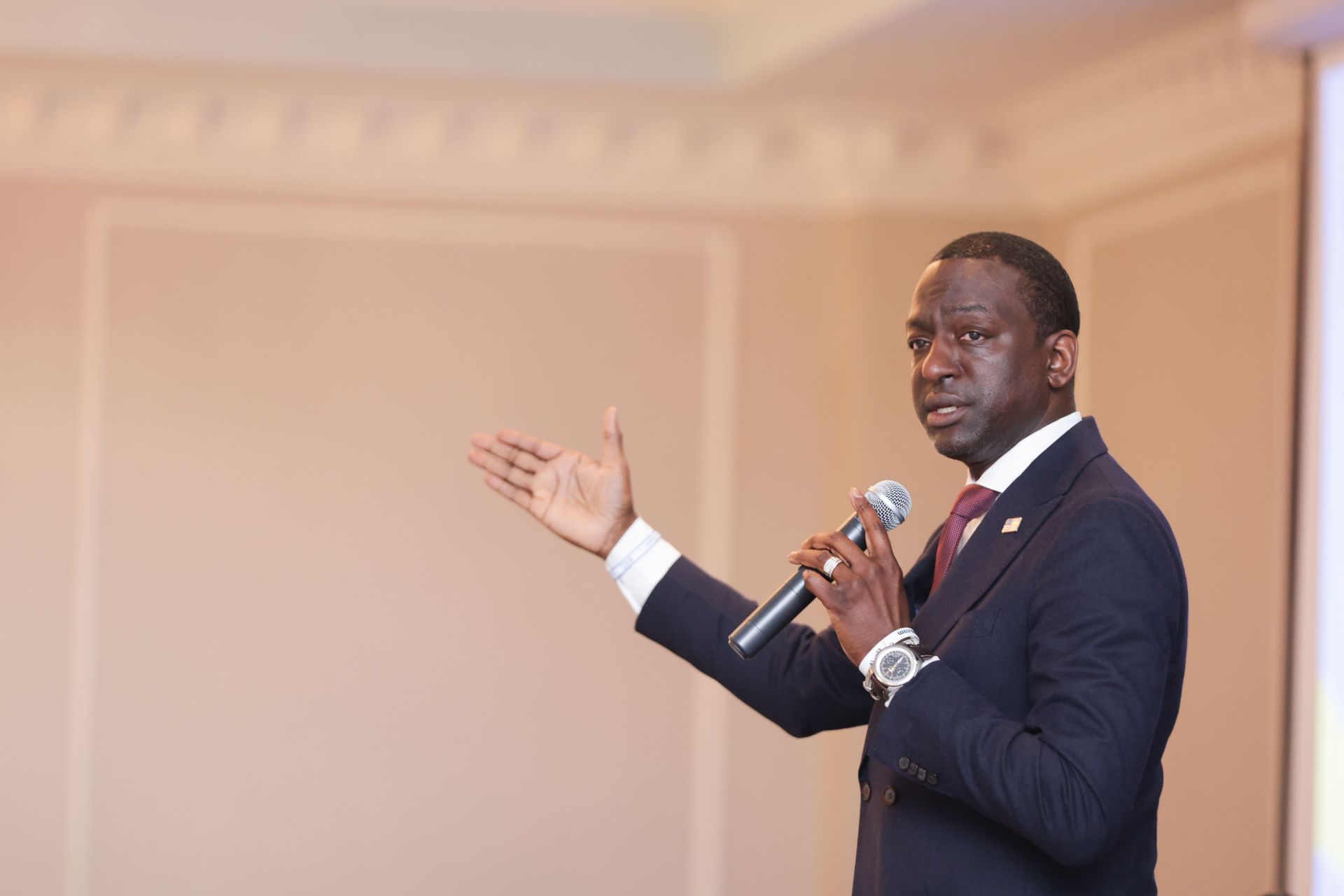 Man in suit speaks into microphone, gesturing with his hand in an event setting.