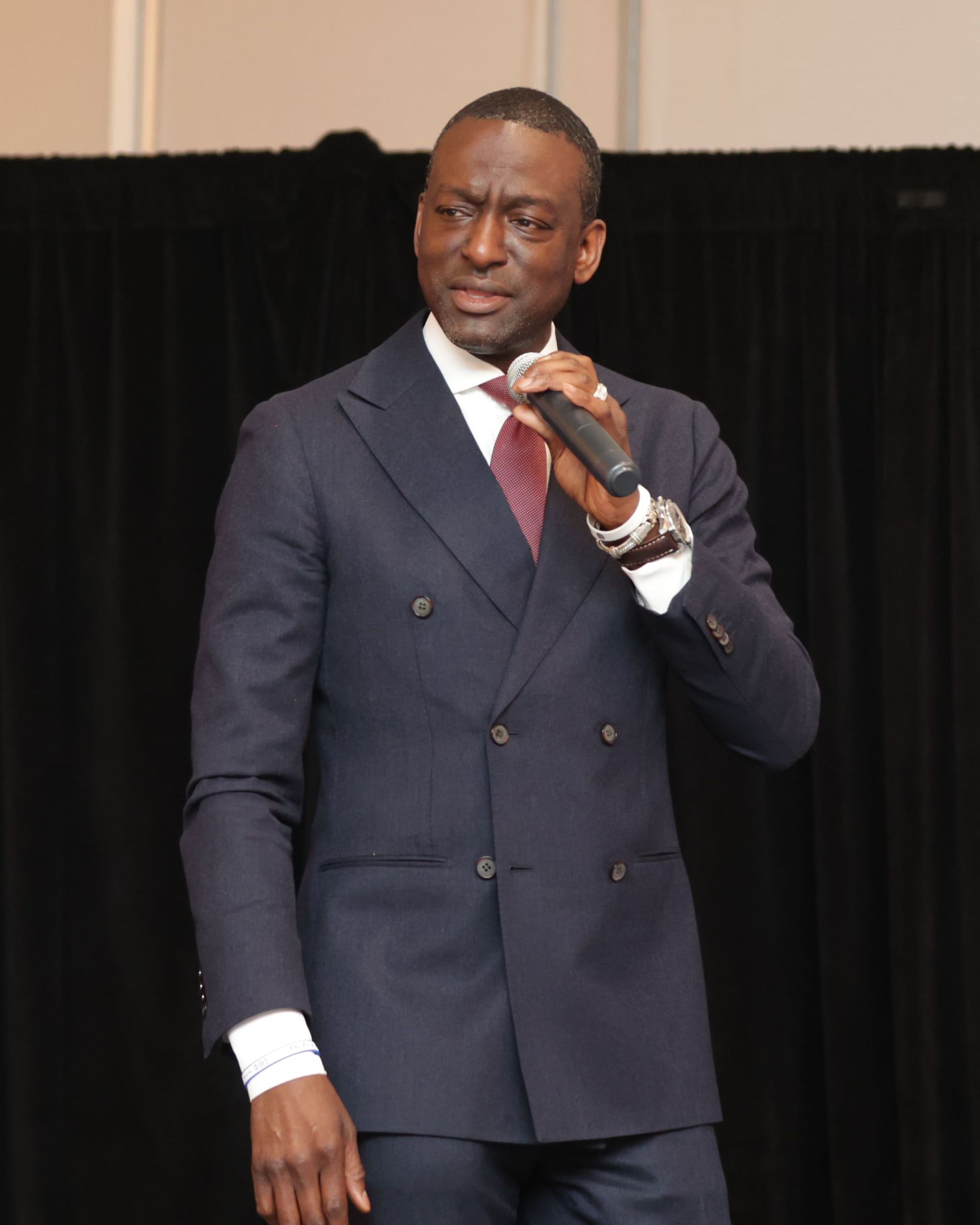 Man in navy suit speaks into a microphone.