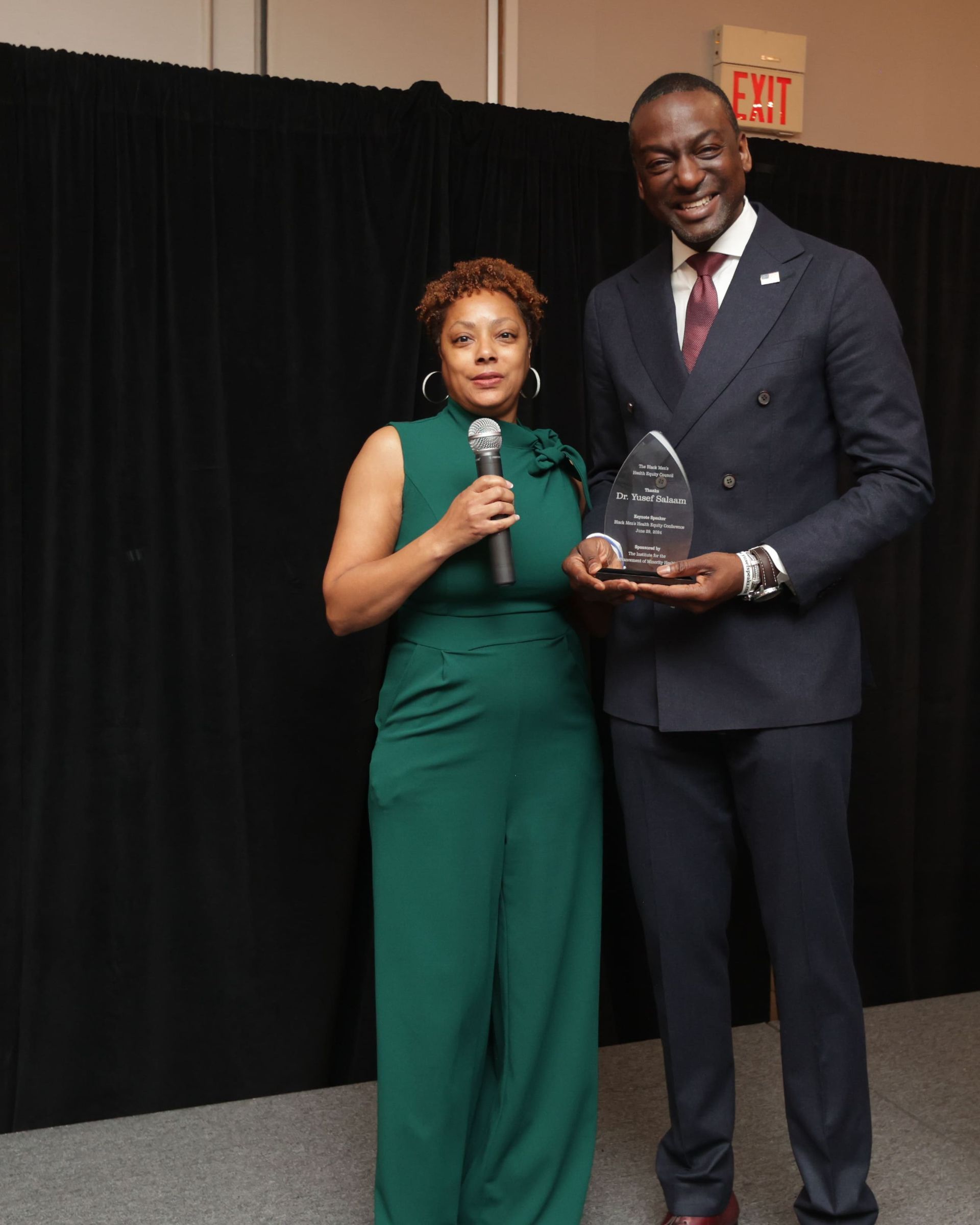 Dr. Melvin | Woman in green jumpsuit holds microphone; man in suit holds award. They stand near a black curtain.