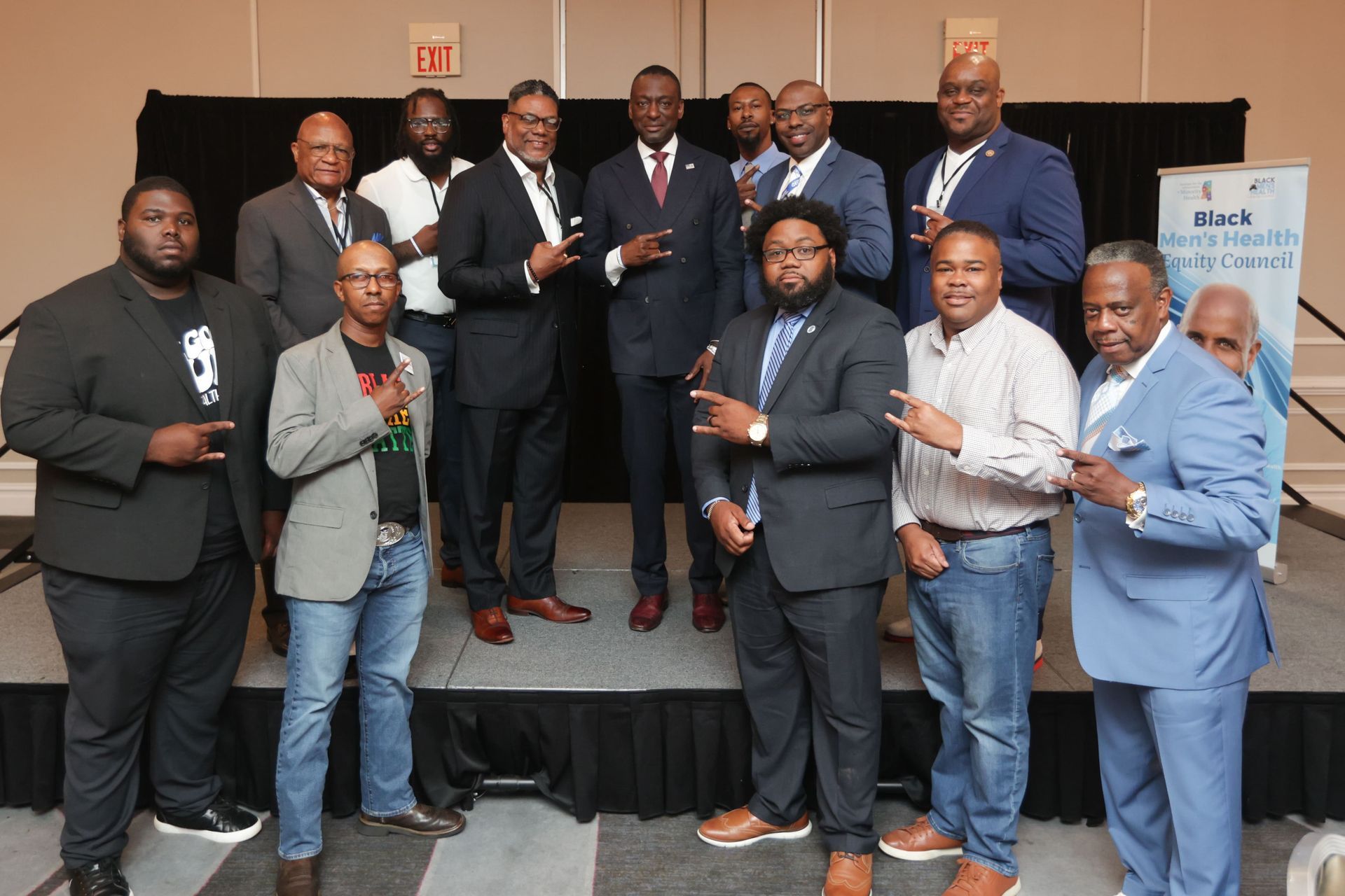 Group of men in suits and casual attire on a stage, some making hand gestures, in a conference setting.