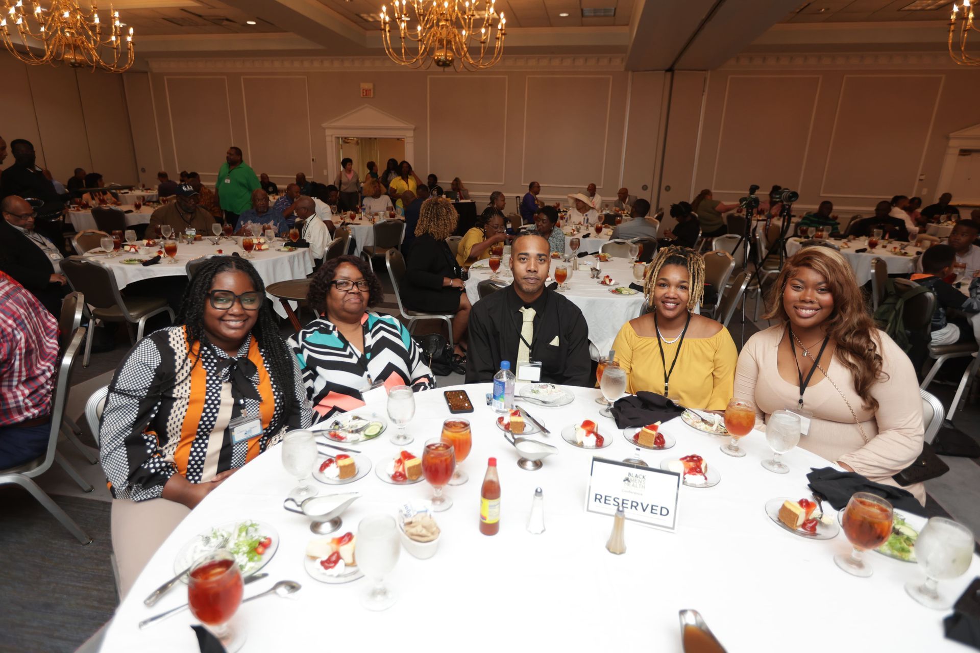 People seated at a round table, posing for photo during a conference or event. Other attendees seated at tables in a large hall.