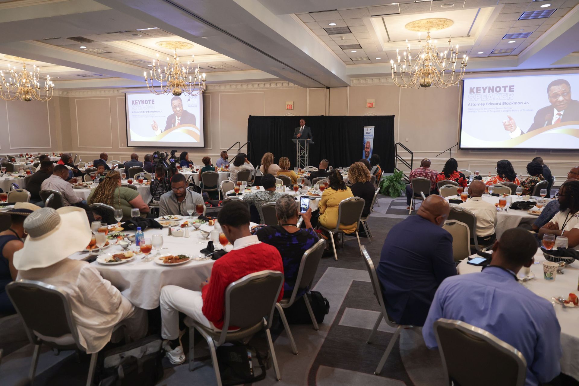 Conference attendees at round tables, listening to a speaker at a podium, with two screens displaying presentations.