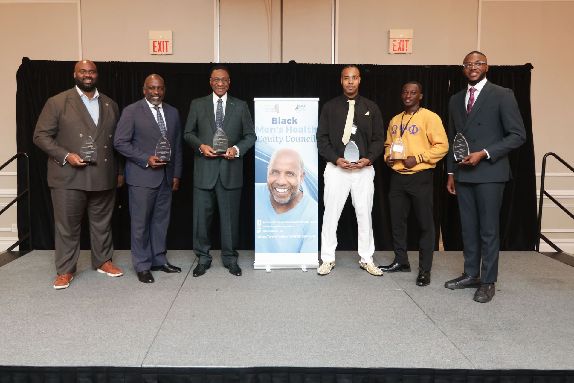 Six people holding awards on a stage with a banner. Men in suits and a casual outfit.