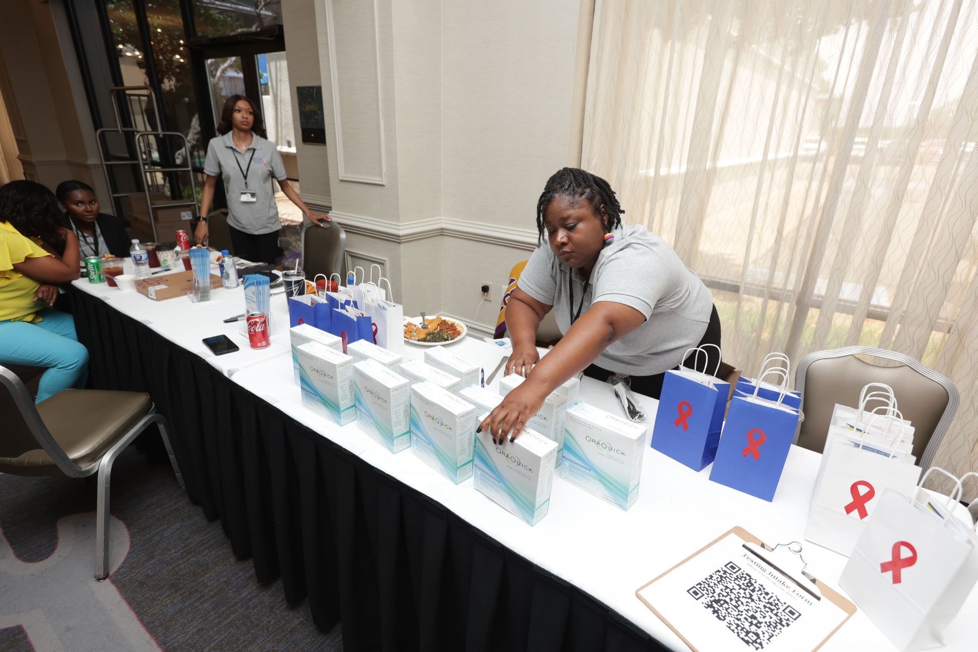 Woman organizing boxes on a table with bags and QR code. Others stand in background at an event.