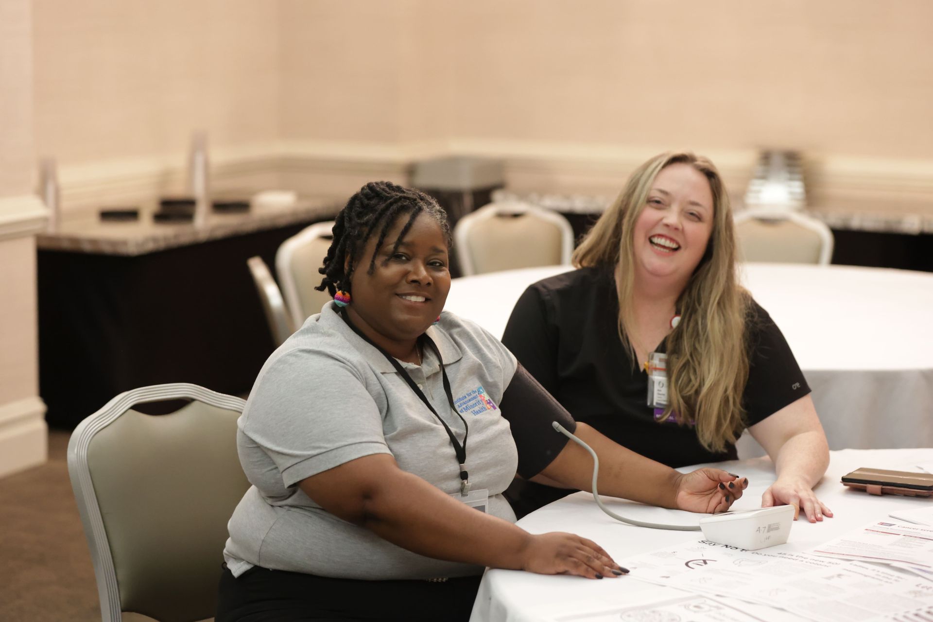 Two smiling women at a table. One has blood pressure cuff on her arm. Meeting setting, white table cloth.