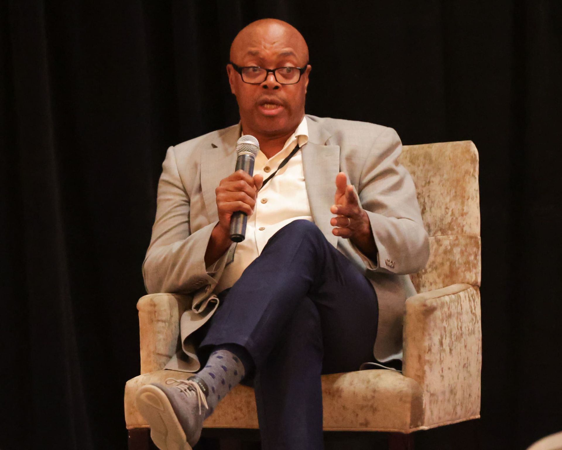 Man in glasses speaks into microphone, seated on chair, gesturing.