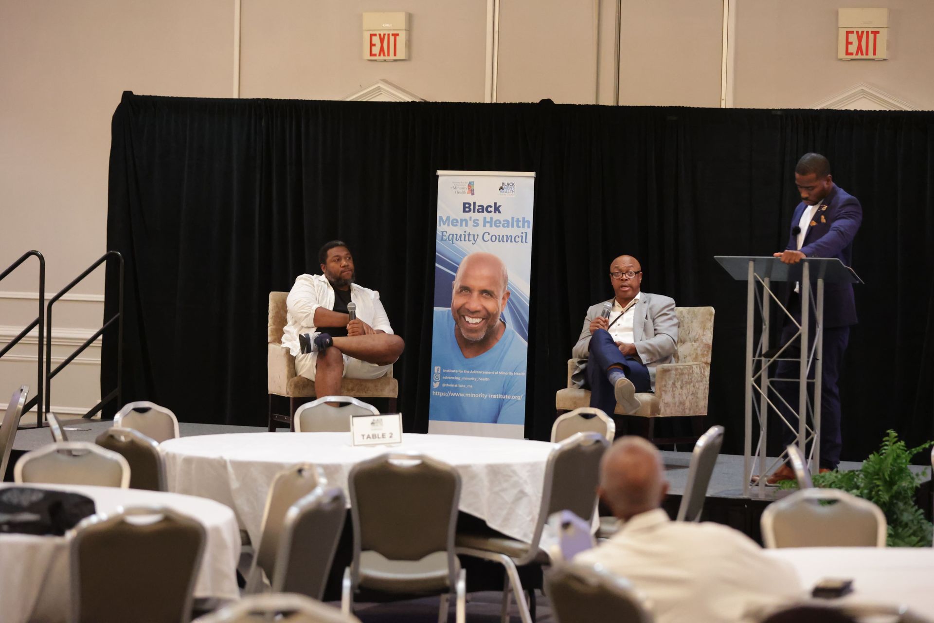 Panel discussion on a stage with a banner. Three men, one at a podium, two seated. Round tables in foreground.