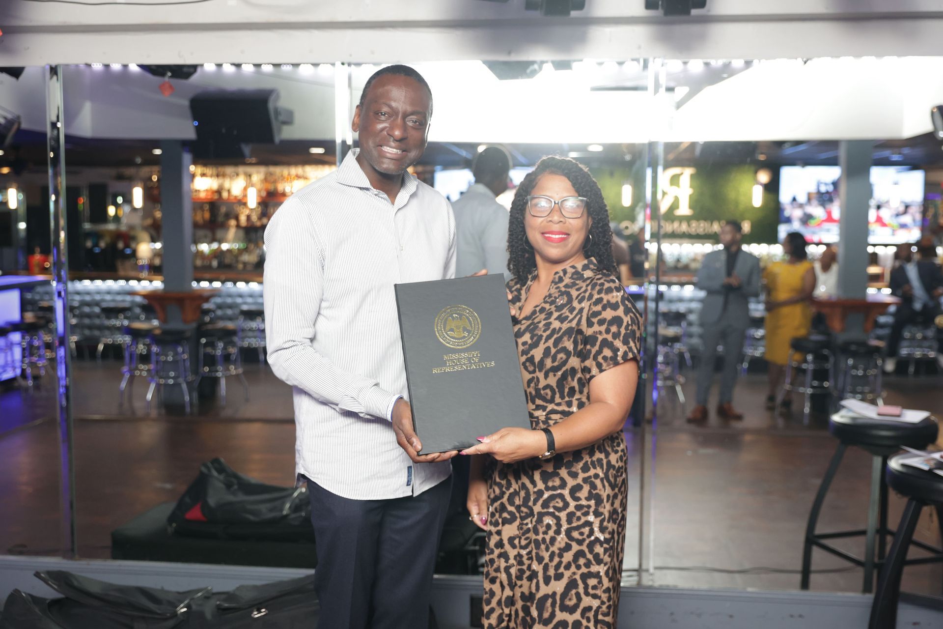 Man and woman holding a framed document inside a bar, smiling. Reflective mirrors behind them.