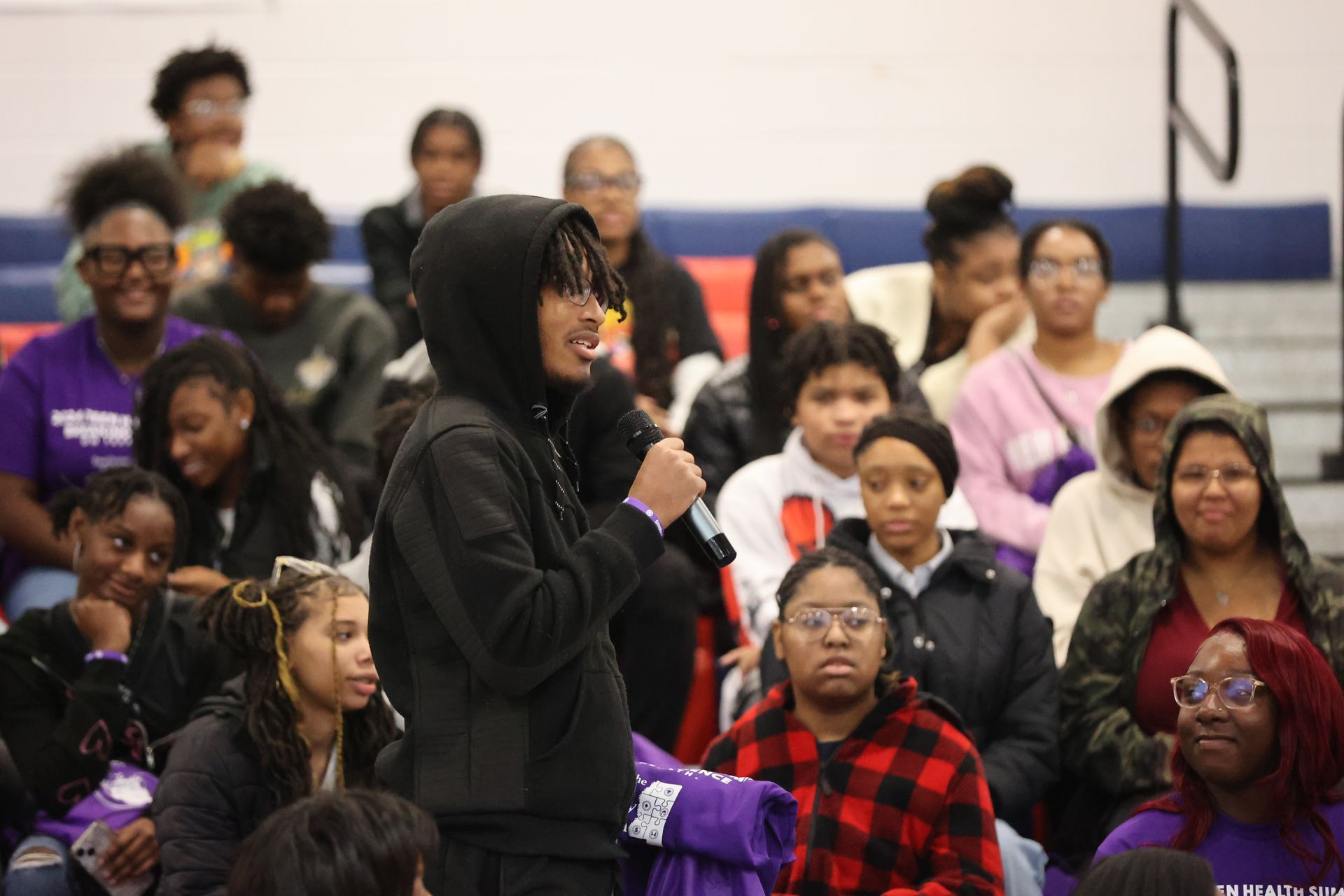 A person speaking into a microphone, standing in front of an audience in a gymnasium.