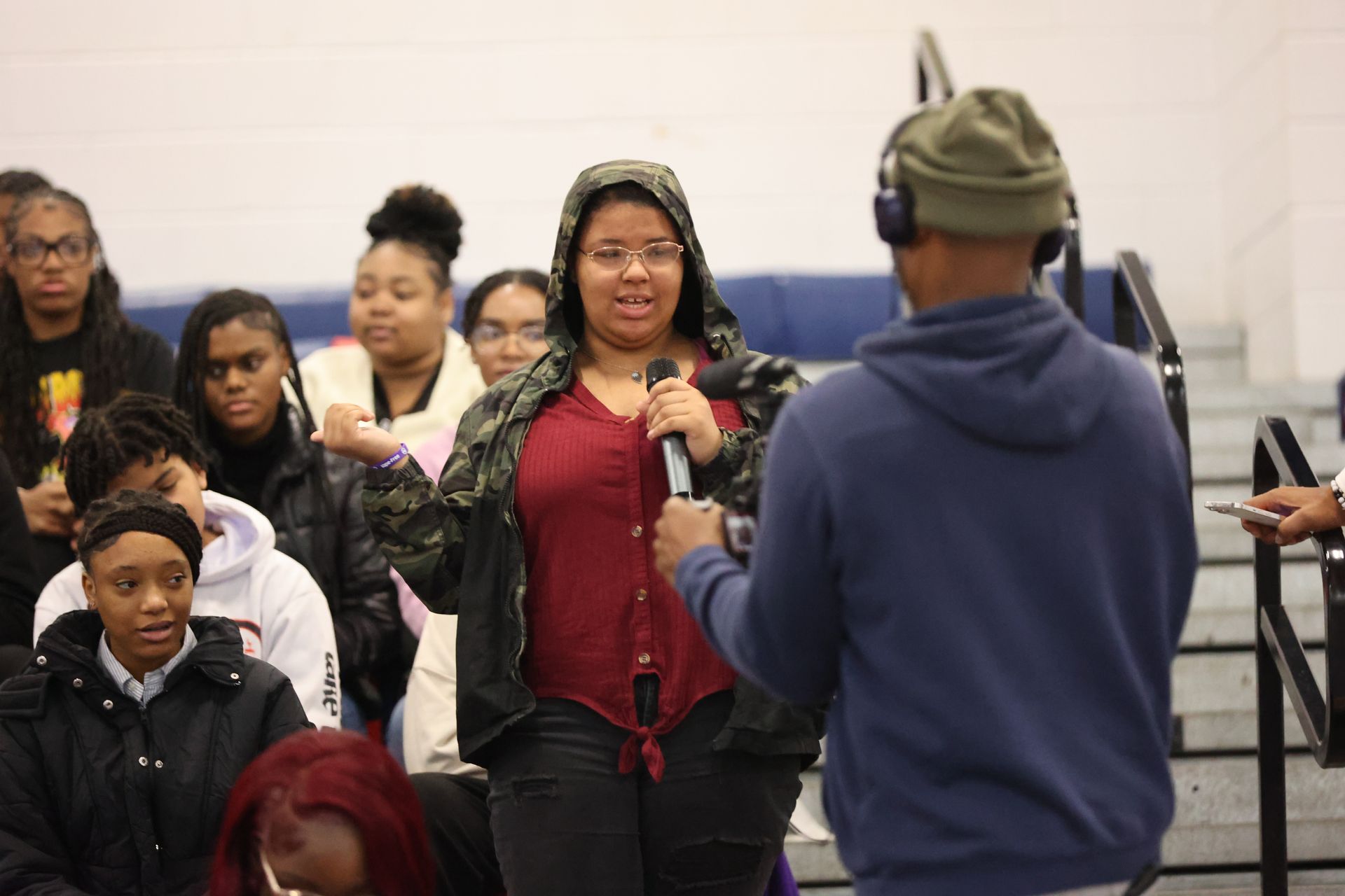A young person speaks into a microphone, addressing a seated audience in a gymnasium.