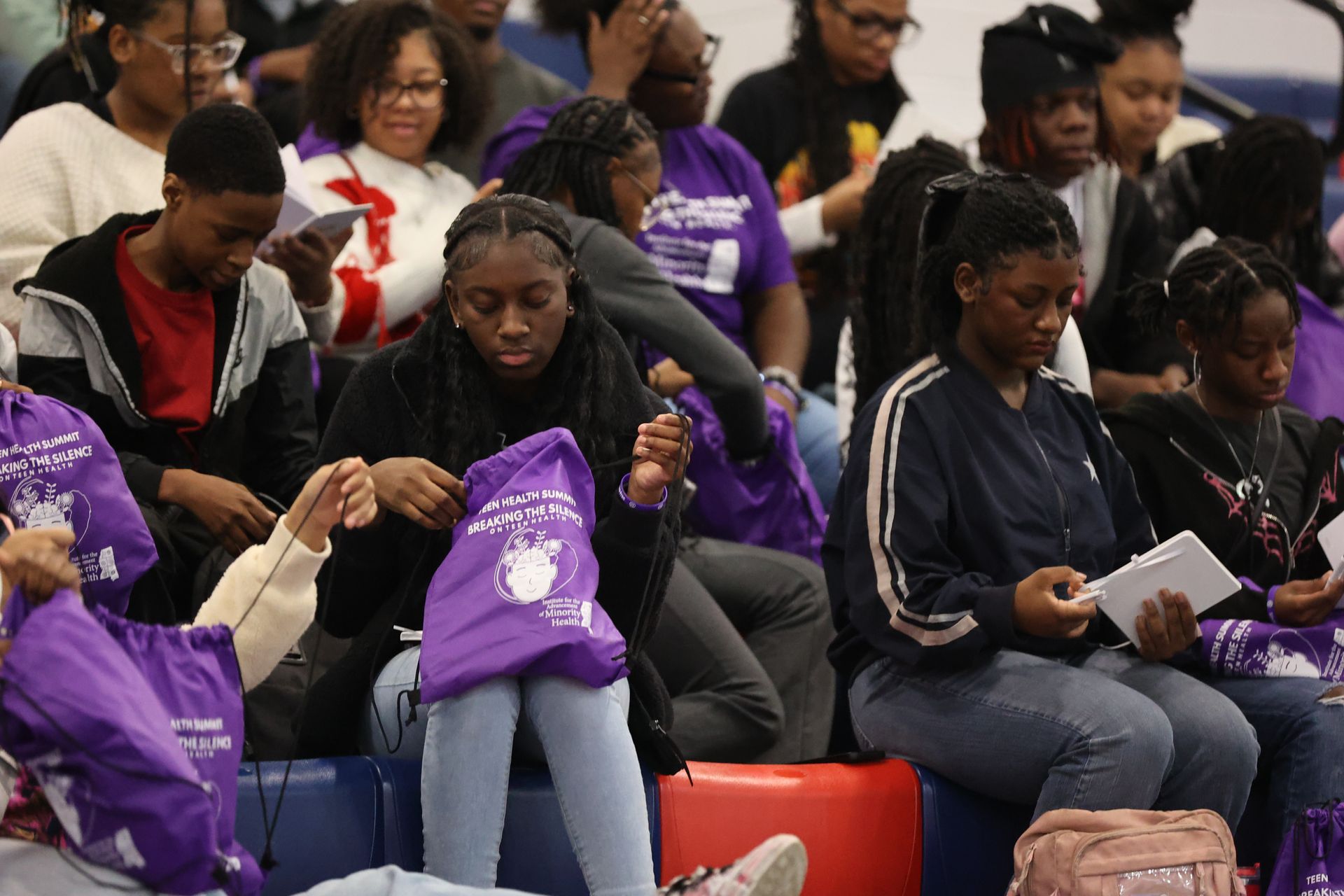People seated at an event, many wearing purple. Some looking down at items, others reading.