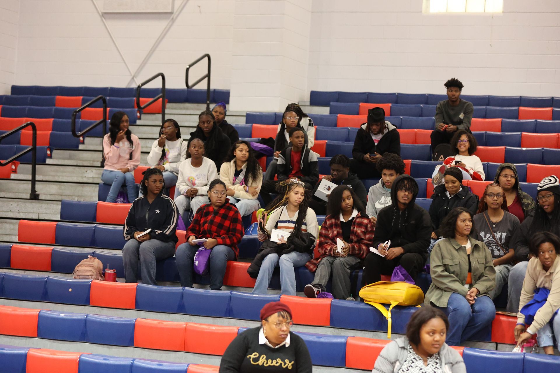People seated in bleachers, attending an event in a gymnasium with red and blue seating.