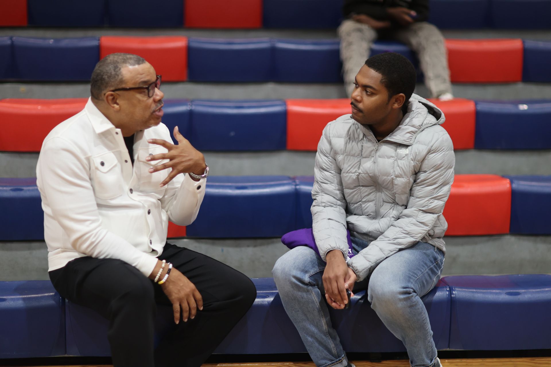 Man in white shirt gestures, talking to person in gray jacket on bleachers.