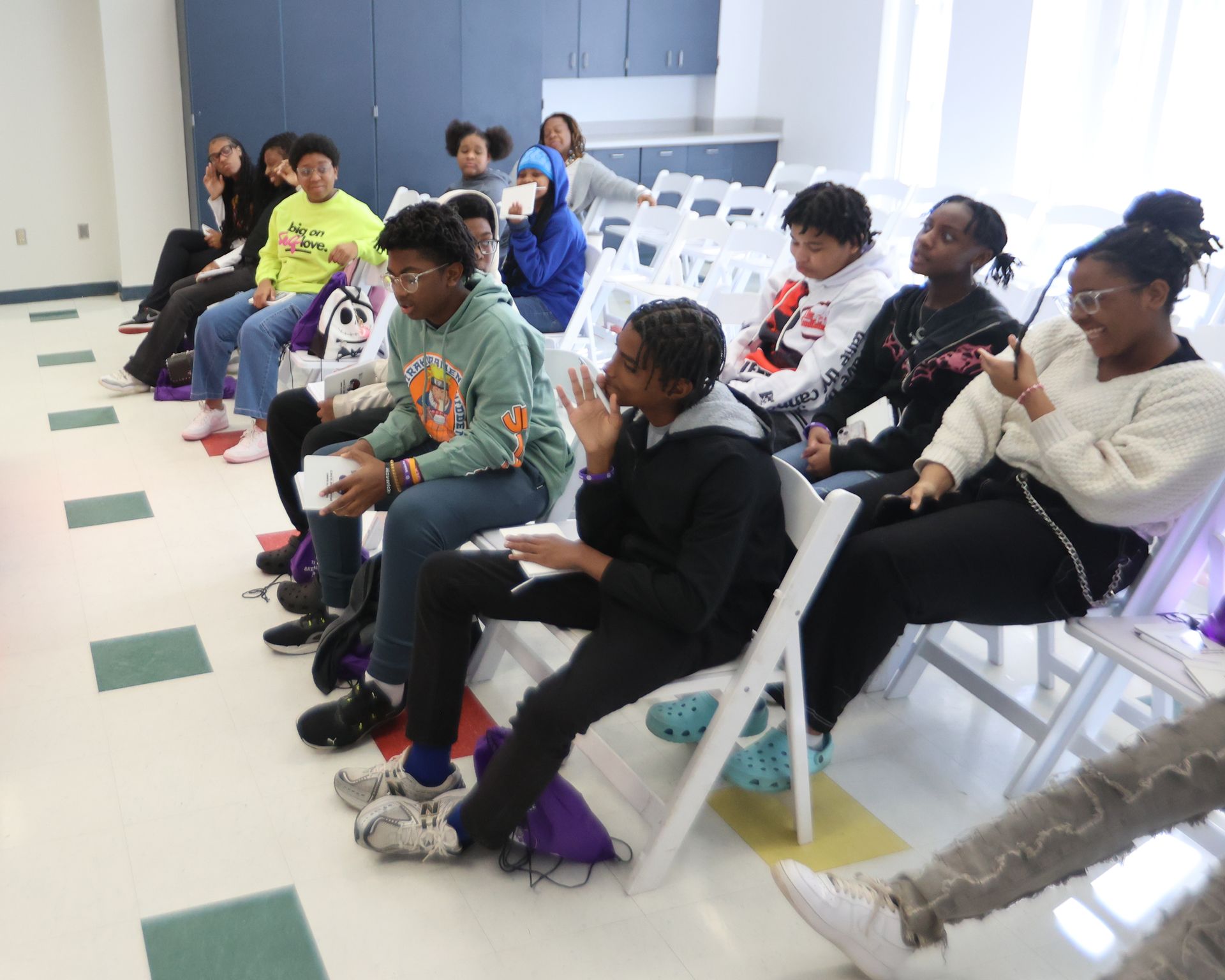 Group of people seated in a room, watching something. Some are gesturing or looking at a device. White chairs arranged in rows.