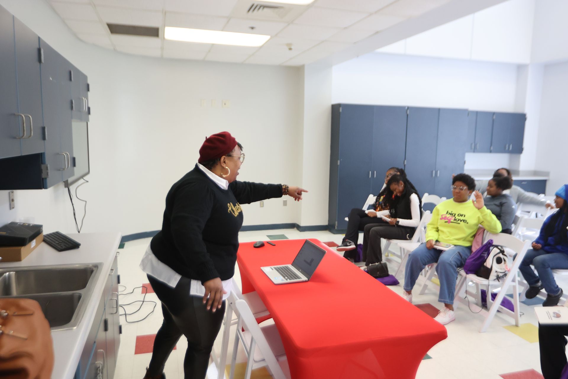 Woman pointing towards seated people in a classroom. Red table with laptop. People are looking at the woman.