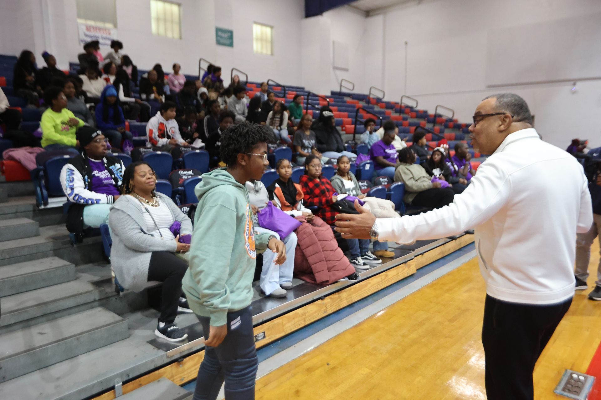 Man shakes hands with a teen in a gymnasium. Spectators in bleachers watch.