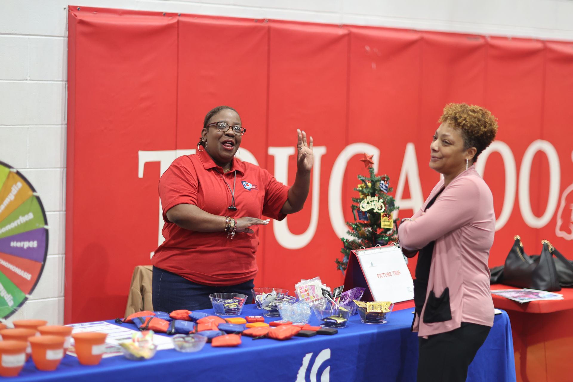 Two people at a booth with red and blue accents. One gestures while the other listens, arms crossed. Dr. Melvin