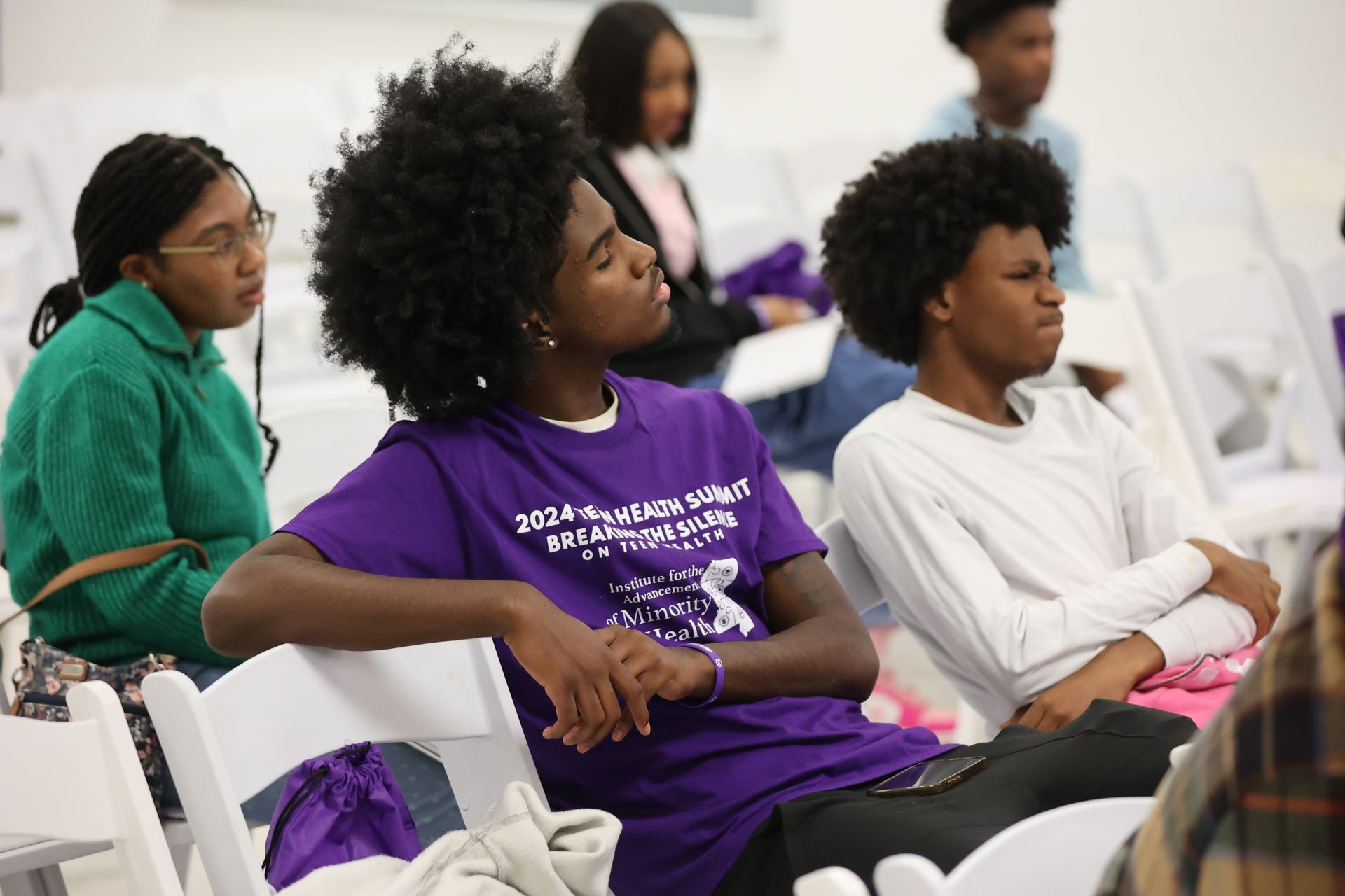 People sitting in a room, listening. One person wears a purple shirt with text and has curly hair.