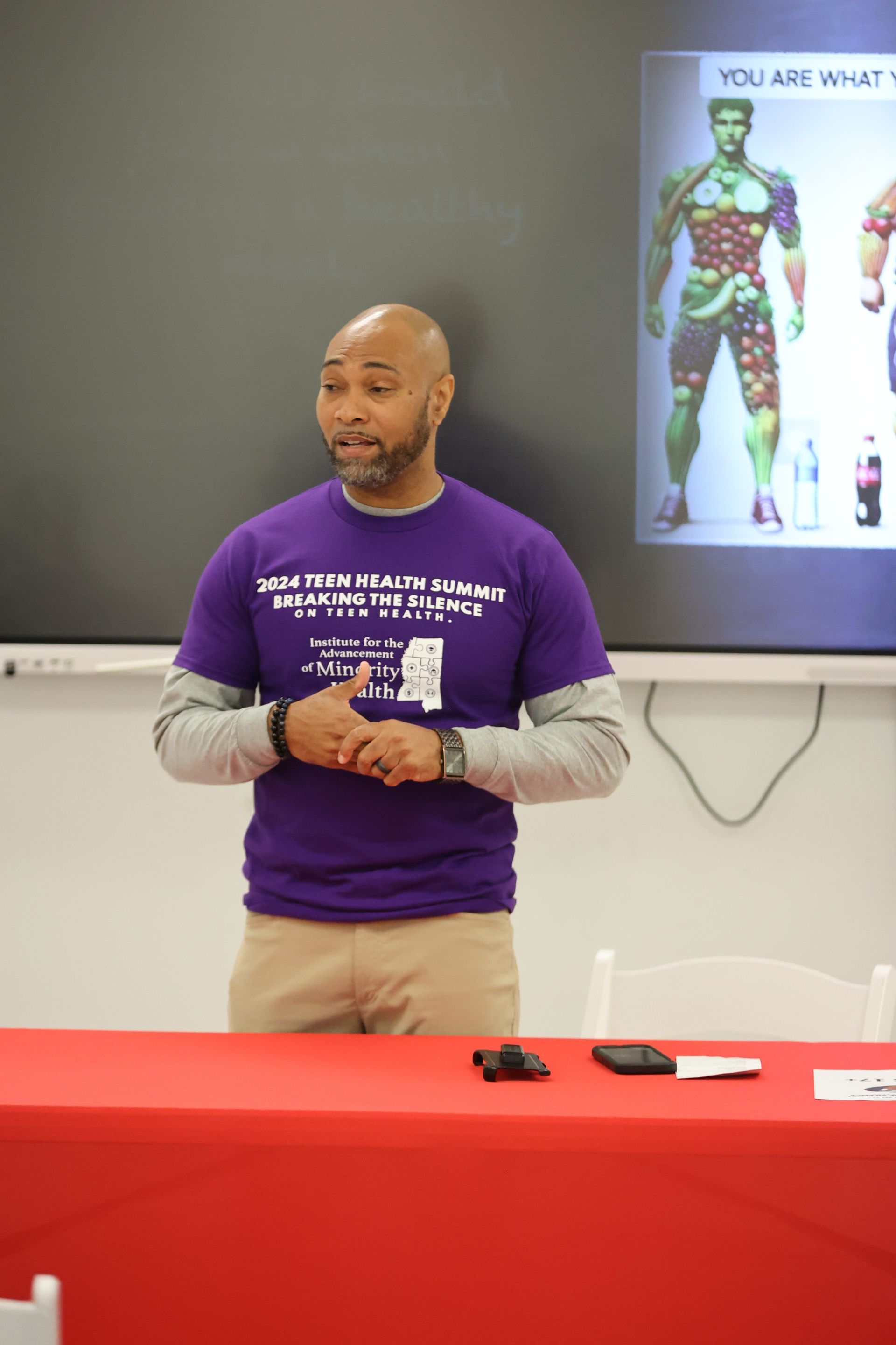 Man speaking at a table, wearing a purple shirt; a health visual is behind him.