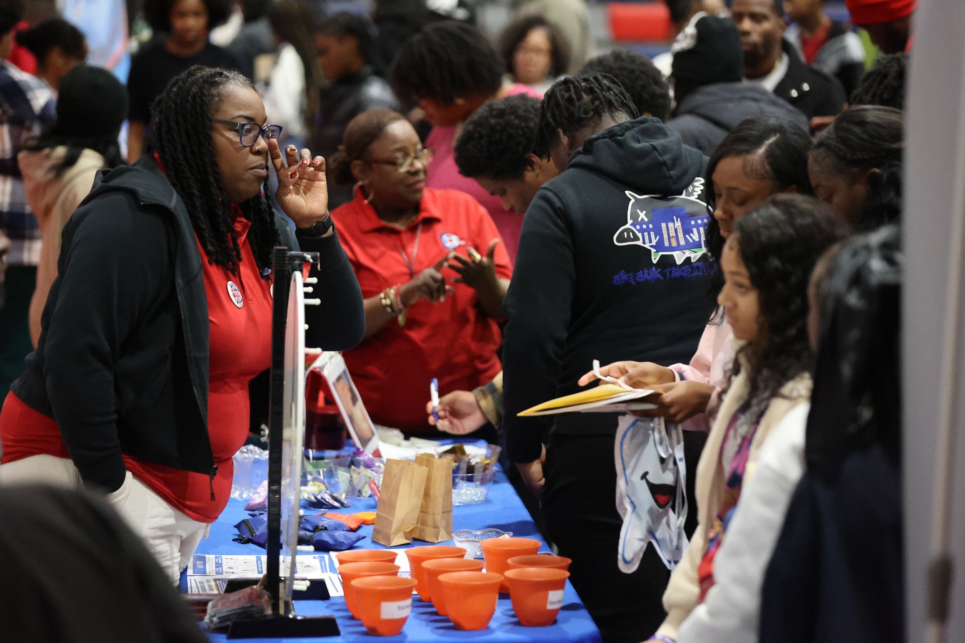 People at a table with brochures and drinks, talking. Woman in red shirt and glasses speaking into a microphone.