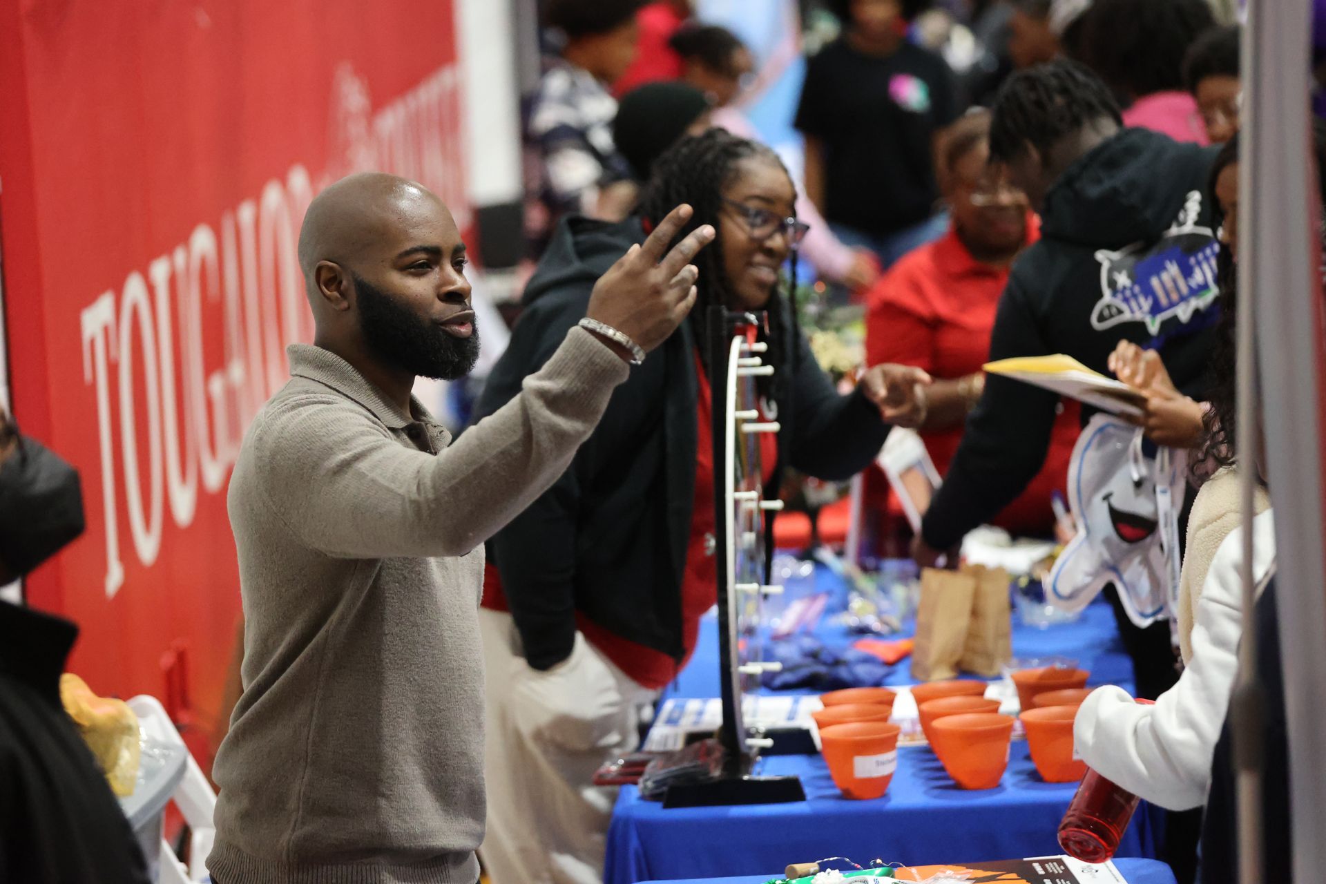 Man gesturing near a table with educational materials; people in the background, likely at a school fair.