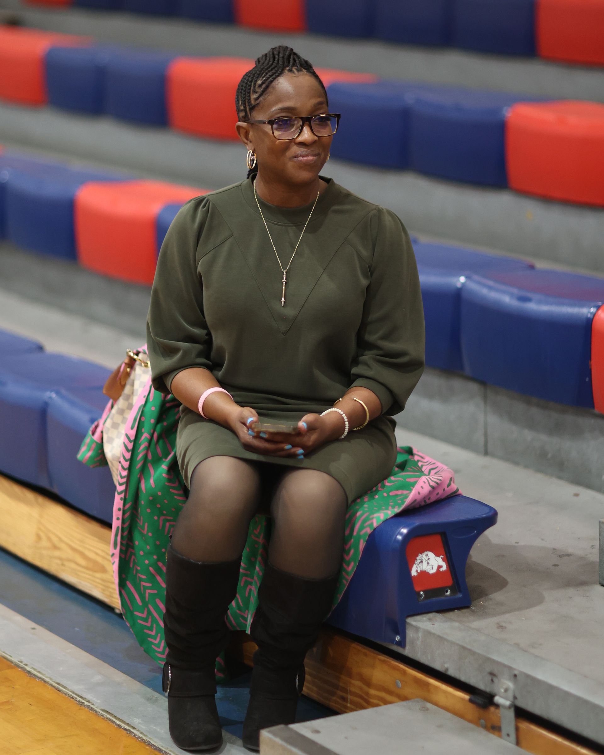 Woman in olive dress and boots sits on bleachers, holding a phone. A blanket rests beside her.