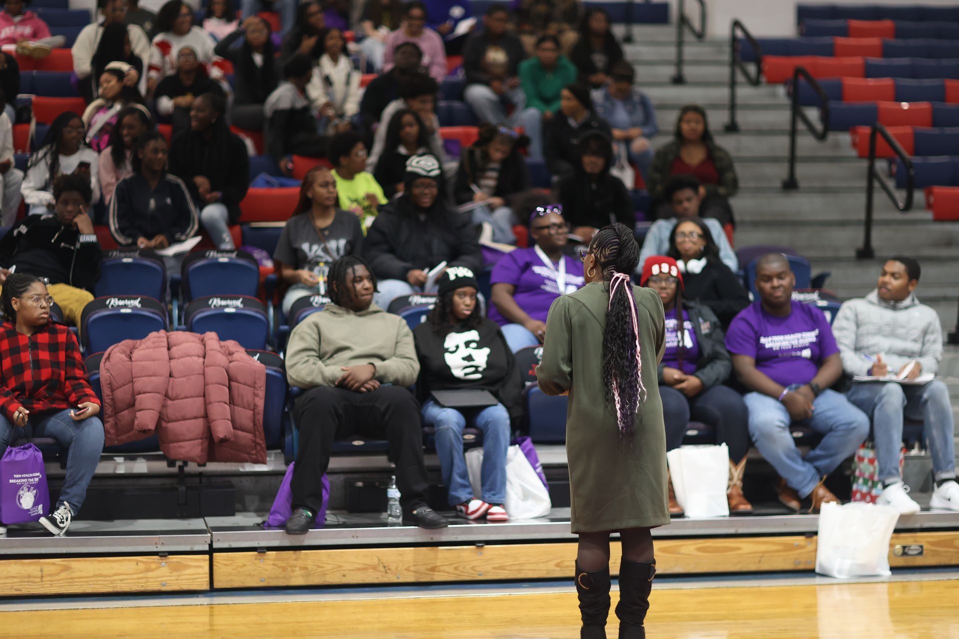 Woman speaking to a seated audience in a gymnasium. People in casual clothes listen, some take notes.