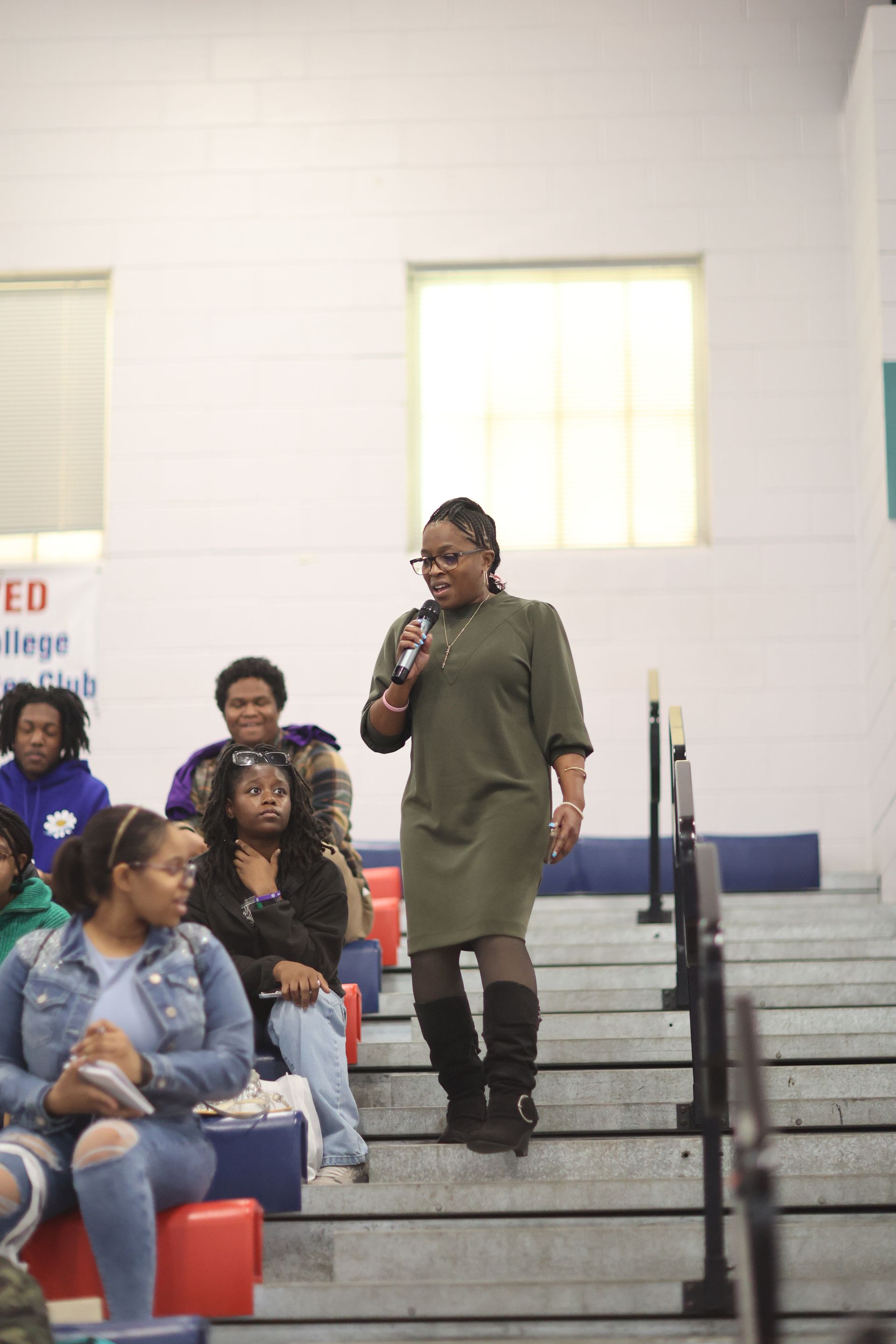 Woman speaking into a microphone, walking down steps in a room with people seated on steps.