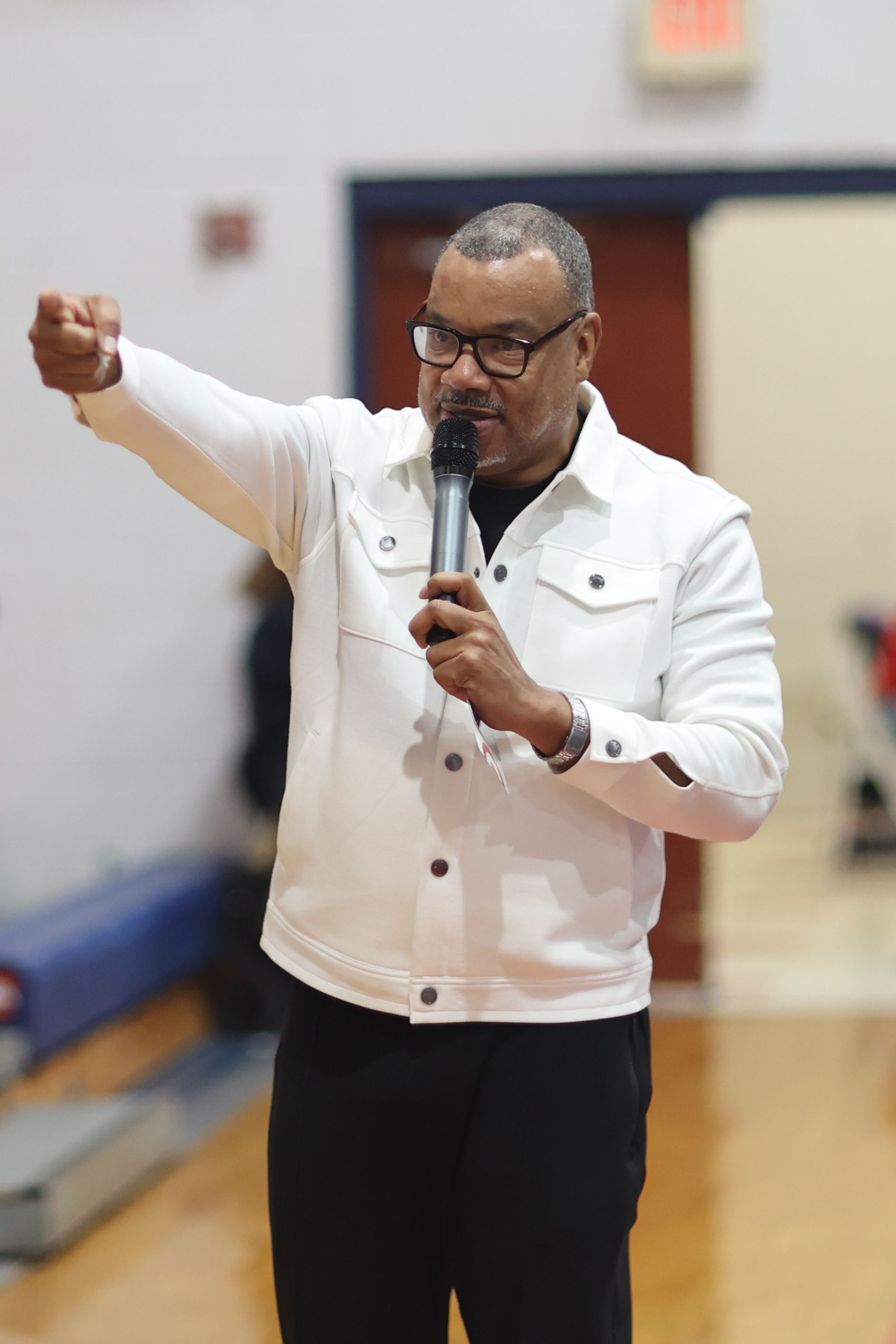 Man in white jacket speaks into microphone, gesturing with his right arm in an indoor setting.