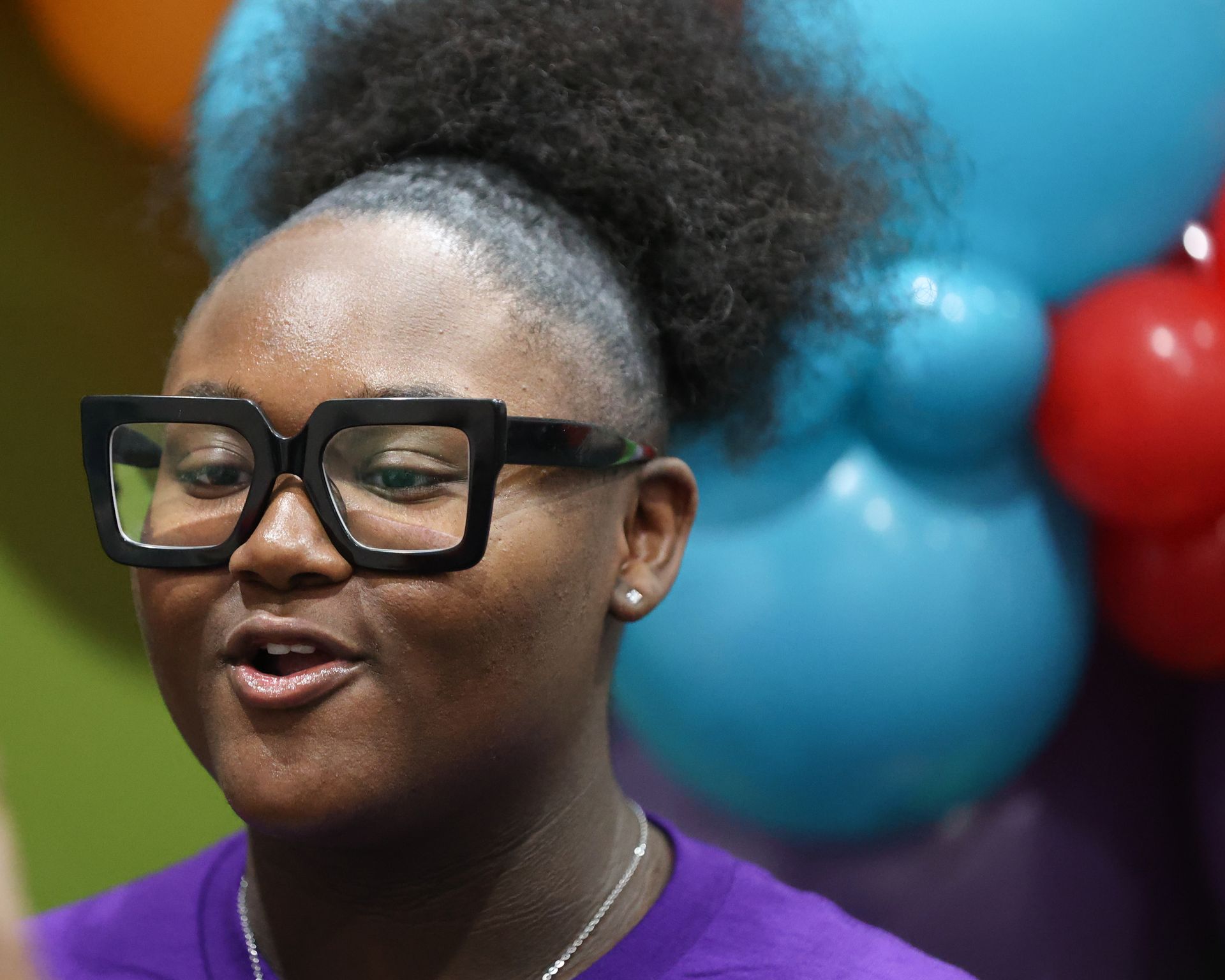 Person with large glasses and afro speaking, near blue and red balloons.